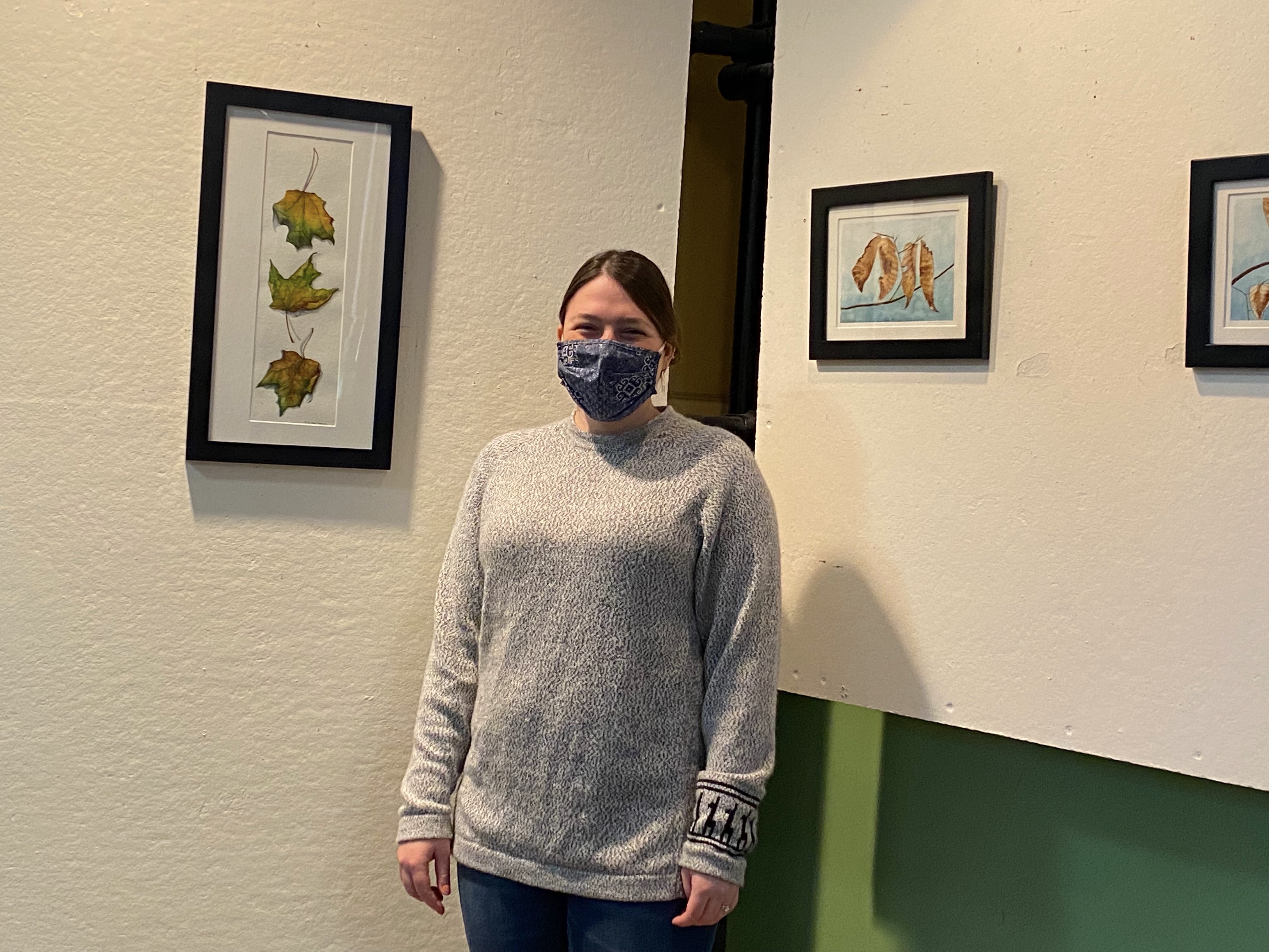 Artist Amy Hook Therrien poses for the camera, between two different display panels each with one painting hung vertically, after installing her artwork in LNT's Gallery space.
